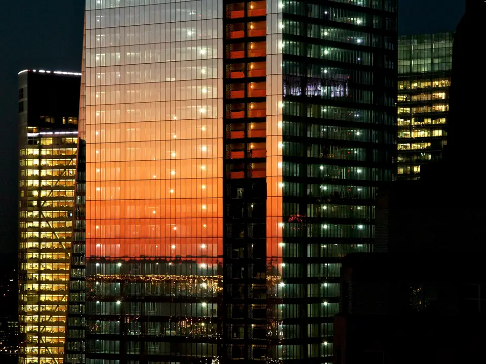 Glass office towers at dusk, the sunset reflected in one facade and the floors lit