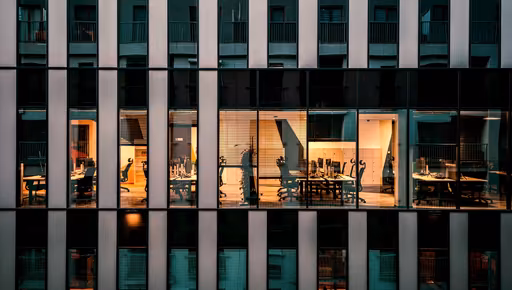 Office facade at blue hour, lit meeting rooms and desks behind the glass